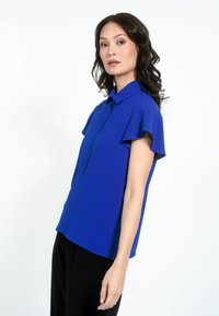 Blå kortærmet blouse med krave, der har flagrende ærmer og en glat tekstur. Båret med sorte bukser mod en hvid baggrund.