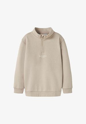 Sweatshirt beige avec un col zippé mi-long, des manches longues et une coupe décontractée. Présente le texte "Édition Studio" sur le devant. Texture de tissu douce.