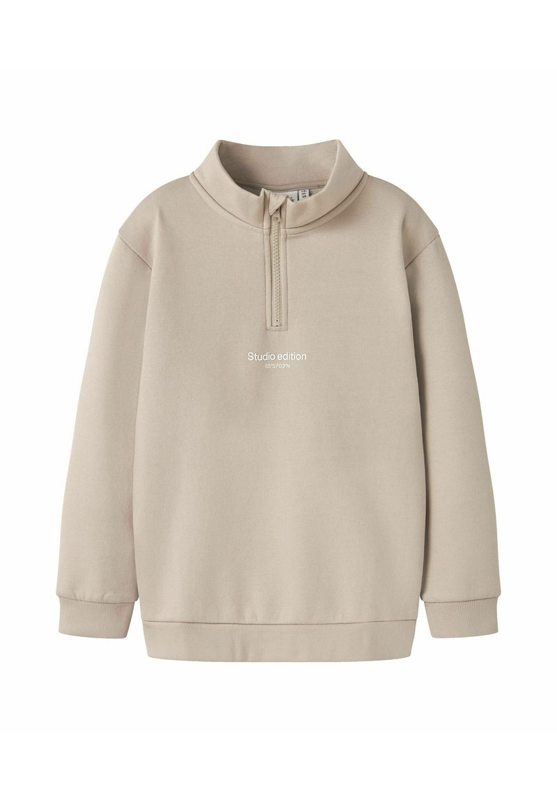 Sudadera beige con cuello de media cremallera, mangas largas y un corte relajado. Presenta el texto "Edición de estudio" en la parte frontal. Textura de tela suave.