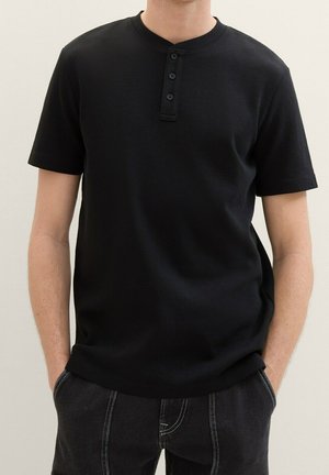Mann trägt ein schwarzes Henley-Shirt mit kurzen Ärmeln und drei Knöpfen, die Hände in den Taschen, kombiniert mit schwarzen Hosen mit weißen Nähten.