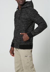 Man som bär en svart mönstrad zip-up huvtröja med händerna i fickorna och beiga cargobyxor med en zip-ficka på sidan.