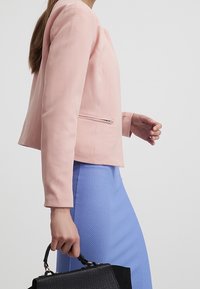 Ljusrosa figursydd jacka med långa ärmar och en dragkedjeficka, i kombination med en strukturerad blå kjol. Hand som håller en svart handväska.