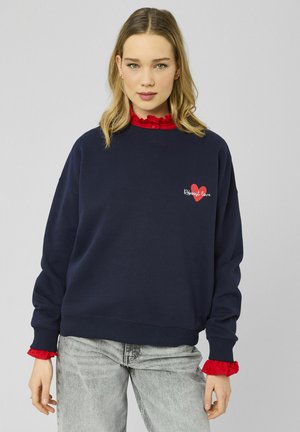Sweat-shirt oversize bleu marine avec poignets côtelés, orné d'un graphique cœur rouge et du texte « Reboot love ». Porté par-dessus un haut rouge à volants.