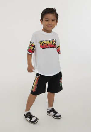 La maglietta bianca presenta una grafica colorata "CALI" sulle maniche; abbinata a pantaloni corti neri con grafiche simili. Il bambino indossa sneaker bianche e nere.