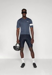 Férfi kerékpáros sötétkék és fekete kerékpáros ruhában, fekete sisakot tartva, fehér zoknival és fekete kerékpáros cipőben, szürke háttér előtt áll.