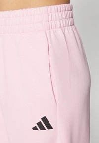 Lys rosa joggebukser med samlet elastisk midje, med sidelommer og en svart trestripe-logo på øvre venstre ben.