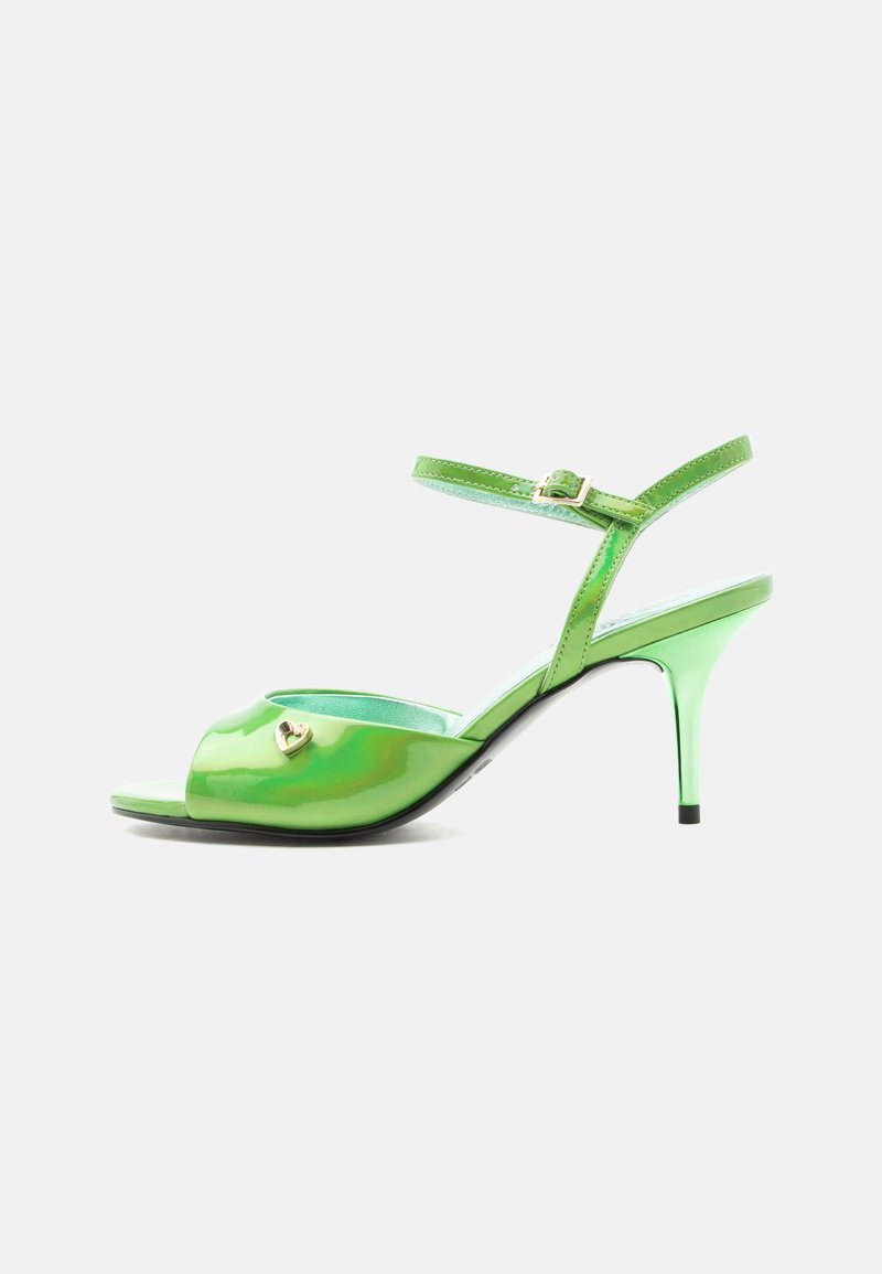 Πράσινο σανδάλι από πατενταρισμένο δέρμα με ανοιχτά δάχτυλα, ψηλό τακούνι, λουράκι στον αστράγαλο και μικρή χρυσή διακόσμηση σε σχήμα καρδιάς στο μπροστινό μέρος.
