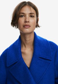 Manteau en mélange de laine bleu avec une finition texturée, grands revers et col cranté. Présente un décolleté en V profond et une coupe décontractée.