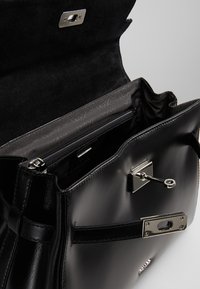 Zwarte leren handtas met een glanzende afwerking, voorzien van zilveren hardware, een interne ritsvak en een gestructureerde voering. Sluiting is metalen.