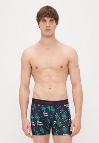 Black boxer briefs with green Christmas tree print and red "JACK & JONES" waistband. Material is stretchy and fitted, with playful holiday graphics.