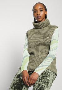 Suéter sem mangas de gola alta em verde oliva, combinado com uma camisa de mangas longas listrada em verde claro e branco e calças de veludo verde escuro.