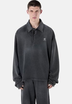 Jonge man die een oversized, verweerd zwart overhemd met lange mouwen en kraag draagt, met een logo en bijpassende losse broek, tegen een witte achtergrond.