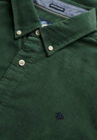 Donkergroen corduroy polo shirt met een button-down kraag, voorzien van lichtgekleurde knopen en een klein blauw embleem op de linkerkant van de borst.