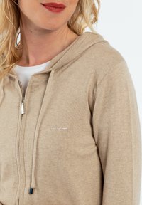 Veste à capuche zippée beige avec une texture douce, capuche à cordon de serrage, poches latérales et logo brodé subtil sur la poitrine.