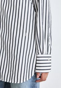 Chemise à manches longues rayée verticalement noir et blanc avec poignets boutonnés, portée avec un jean bleu clair.