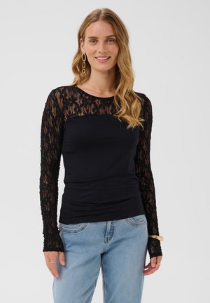 Femme aux cheveux blonds ondulés porte un top en dentelle noire à manches longues et un jean bleu clair, souriante, avec des boucles d’oreilles et un bracelet en or.