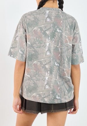 Person med fletninger iført en løs grå t-shirt med camouflage mønster og en sort denim mini nederdel med frynset kant, vendt væk.