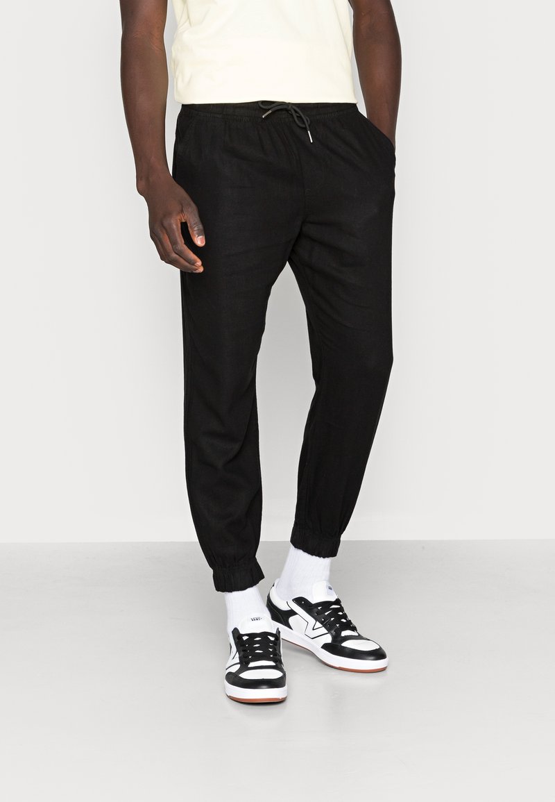 Zwarte tapered joggers met elastische boorden, een trekkoord in de taille en een lichtgewicht stof, gecombineerd met zwart-witte sneakers en witte sokken.