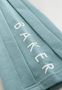 Svetlomodrá látka s bielymi písmenami, ktoré diagonálne po šitých švoch tvoria slovo „BAKER“.