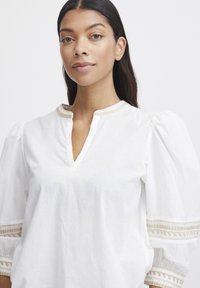 Blouse blanche avec un décolleté en V, des manches bouffantes et des détails en dentelle élaborés sur les poignets. Fabriquée en tissu léger avec une finition texturée.