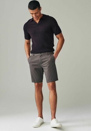 Temno srajčno polo s kratkimi rokavi in žepnim dekoltejem, v kombinaciji s sivimi hlačami. Obut v bele superge, kar prikazuje sproščen, prilega joč slog.