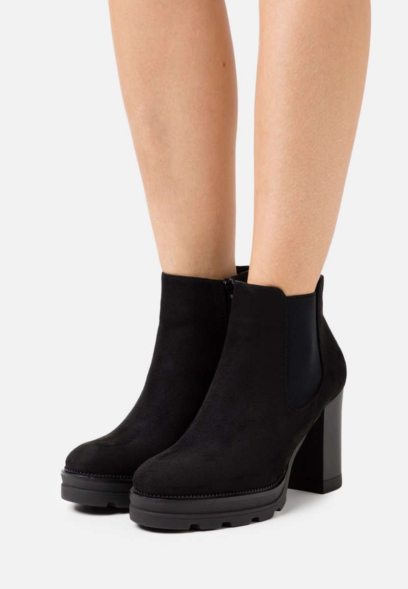 Bottines en daim noir avec talons hauts épais et panneaux élastiques latéraux portées sur des jambes nues, sur fond blanc.