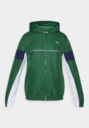 Grønn sportsjakke med hette, med marineblå og hvite ermer, glidelåslukking og en liten logo på brystet. Glatt stofftekstur.