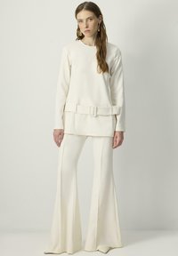 Kreem värvi, pika varrukaga topp, millel on vöödiga vöökoht ja laienevad, samas toonis püksid. Sile tekstuur, minimalistlik disain ja voolujooneline siluett.