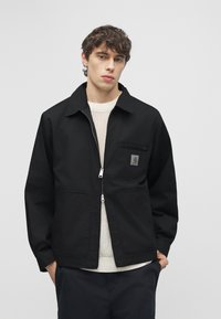 Νέος άνδρας που φοράει μαύρο τζάκετ με φερμουάρ πάνω από ένα λευκό πλεκτό πουλόβερ, στέκεται με τα χέρια στις τσέπες μπροστά σε ένα απλό γκρι φόντο.