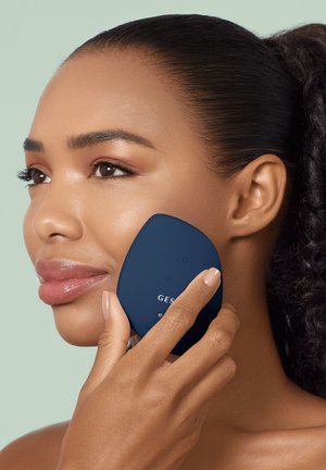 Femme utilisant un appareil facial portatif bleu foncé sur sa joue, démontrant un soin de la peau ou un traitement de beauté.