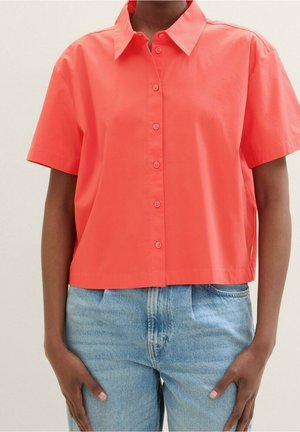 Persona con una camisa de manga corta color coral brillante abotonada, combinada con jeans de mezclilla azul claro de cintura alta, de pie con las manos a los lados.