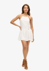 Witte mouwloze jurk met kantstructuur bij de bovenkant en een uitlopende, gelaagde rok. Gecombineerd met bruine veter sandalen.