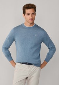 Blauwe crewneck trui gemaakt van zachte stof, met geribbelde manchetten en halslijn, en een klein geborduurd logo op de borst. Gecombineerd met lichte broek.