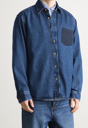 Personne portant une chemise en jean bleue à boutons avec une poche foncée contrastante sur une chemise rayée à col et un jean bleu.