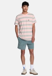 T-shirt en coton rayé rose, vert et bleu, avec des manches courtes et une poche sur la poitrine. Associé à un short bleu clair et des baskets.