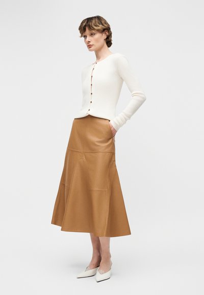Γυναίκα με κοντά μαλλιά που φοράει λευκό πουλόβερ με κουμπιά, καφέ midi δερμάτινη φούστα και λευκά παπούτσια με μυτερή μύτη, στέκεται με τα χέρια στις τσέπες.