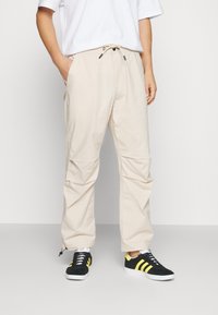 Beige Cargohosen mit Kordelzug am Bund, Seitentaschen und elastischen Bündchen. Kombiniert mit schwarzen Sneakern mit gelben Akzenten.