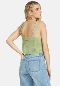 Top corto lavorato a maglia verde chiaro con bretelle larghe e motivo testurizzato, abbinato a jeans chiari a vita alta. La vista posteriore mostra un design aperto.