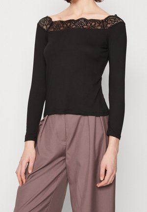 Kvinde iført sort langærmet off-shoulder top med blonderkant og løse malva højtaljede bukser mod en ensfarvet baggrund.