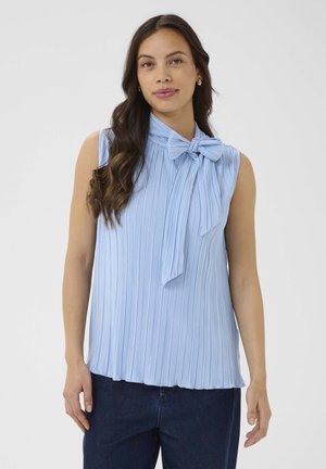 Žena s dlhými tmavými vlasmi, oblečená v bezrukávovej svetlomodrej plisovanej blúzke s veľkou mašľou, v kombinácii s tmavomodrými džínmi.