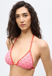 Γυναίκα με σγουρά σκουρόχρωμα μαλλιά, φορούν ροζ και κόκκινο μπικίνι τοπ σε σχήμα τρίγωνου, σε απλό φωτεινό φόντο.