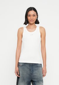 Άσπρο εφαρμοστό τοπ χωρίς μανίκια με ριμπ υφή και στρογγυλή λαιμόκοψη, με ένα μικρό ασημένιο λογότυπο στο κέντρο.