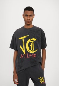 Tmavě šedé oversized tričko se silným žlutým a červeným grafickým textem "JC MILAN" a akcenty v leopardím vzoru. Tkanina vypadá měkce a uvolněně.
