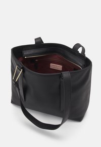 Sac fourre-tout en cuir noir avec une surface texturée. L'intérieur présente un revêtement bordeaux, une poche zippée et une étiquette en cuir de marque. Deux poignées.