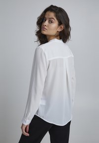 White, sheer long-sleeve blouse with a buttoned cuff and back pleat detail, paired with black trousers. Smooth texture, simple design.