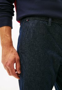 Donkerdenim broek met een gestructureerde afwerking, voorzien van een voorzak en een rood-wit label zichtbaar aan de tailleband.