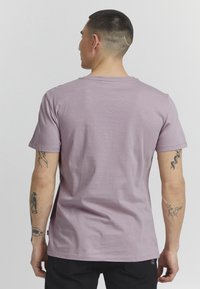 Kortermet t-skjorte i lavendel stoff med rund hals. Har glatt tekstur og synlig tatovering på armen. Svart merkelapp ved kanten.
