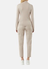 Beige Strick-Turtleneck-Pullover kombiniert mit figurbetonten beigen Cargo-Hosen aus Kunstleder, ausgestattet mit seitlichen Taschen und weißen Turnschuhen.