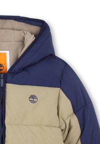 Giacca imbottita con una parte superiore blu navy e una parte inferiore beige. Presenta un cappuccio, una zip frontale e un logo sul petto. Texture morbida.