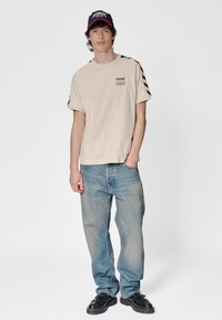 Beige T-shirt met korte mouwen, voorzien van een zwart logo en gestreepte accenten, gecombineerd met lichtblauwe spijkerbroeken en zwarte veterschoenen, gedragen met een zwart en rode pet.